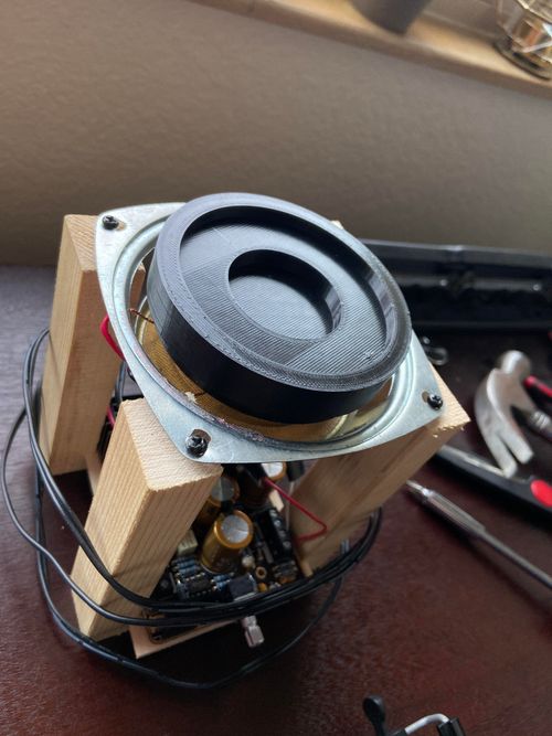 A top front shot of a speaker, with a 3D printed dish on top, bolted onto 4 pine wood posts. Underneath the speaker is mounted a motherboard. Wrapped around the posts is a wire that connects to a wall plug.